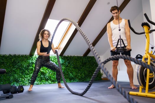 Full body of young sporty woman in activewear exercising with battle ropes during training near fitness buddy in contemporary gym with equipment