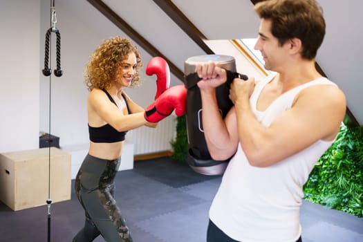 Cheerful powerful young female boxer in red boxing gloves punching bag while practicing with male instructor in gym