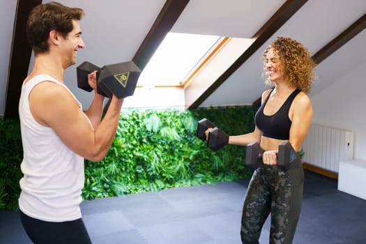 Smiling sporty woman and man lifting heavy dumbbells while exercising in gym with pitched roof during intense training