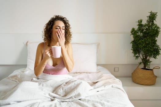 Young woman in nightwear yawning while holding coffee mug in bed during morning
