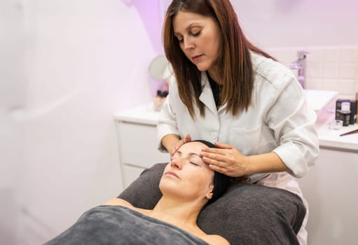 Concentrated adult female cosmetician in white uniform sitting and gently massaging forehead of customer during skincare routine in beauty salon
