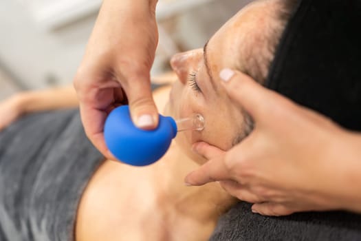 From above of hand applying cupping therapy with blue facial cup to woman with dark hair during skin treatment in beauty spa