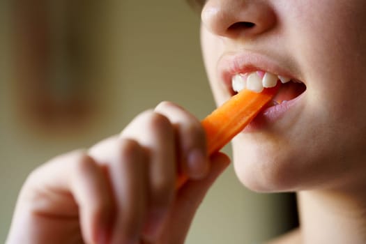 Closeup of crop unrecognizable teenage girl eating fresh healthy carrot slice at home