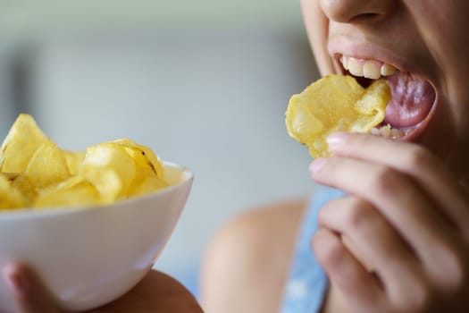 Crop anonymous teenage girl with mouth open about to eat crispy potato chip while holding bowl at home