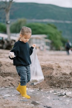 Little girl in rubber boots and gloves with a bag walks through shallow water looking for garbage. High quality photo