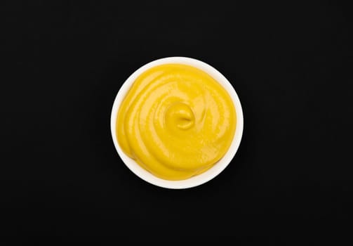 Close up one white ceramic bowl of yellow American mustard on black table background with copy space, top view, directly above
