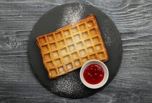 Close up big sweet traditional American or Belgian Brussels waffle with powder sugar and raspberry jam served on black slate board on table, elevated top view, directly above