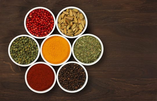 Group of white ceramic bowls with assorted spices over dark wooden table background with copy space, elevated top view, directly above