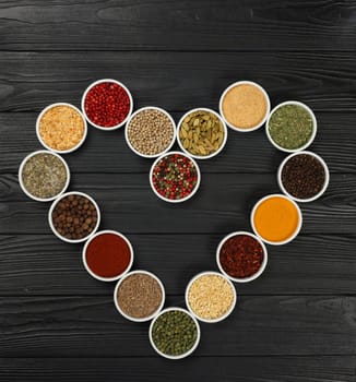 Heart shaped frame of assorted spices in white ceramic bowls over dark wooden table background with copy space, elevated top view, directly above