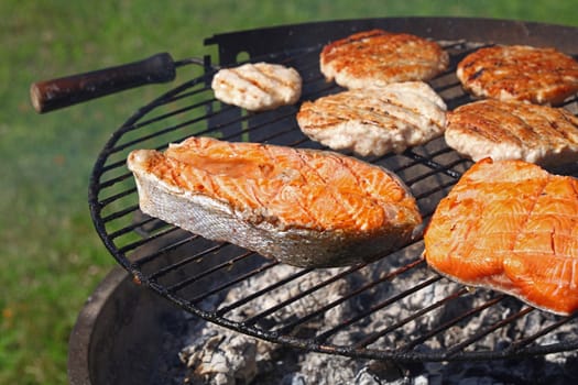 Close up chicken or turkey meat burgers for hamburger and salmon fish steak prepared on outdoor bbq grill, high angle view