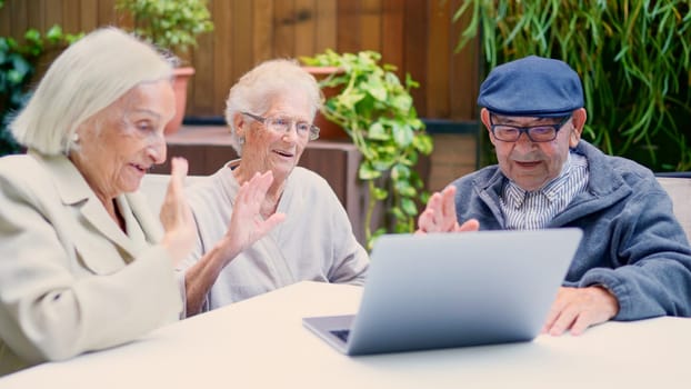 Three seniors using laptop and waving during a video call outdoors in a nursing home