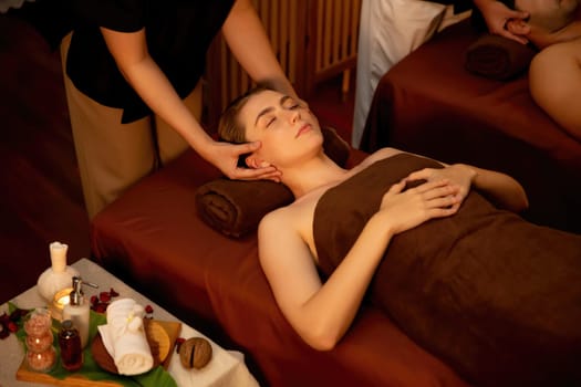 Couple customer enjoying relaxing anti-stress head massage and pampering facial beauty skin recreation leisure in warm candle lighting ambient salon spa in luxury resort or hotel. Quiescent
