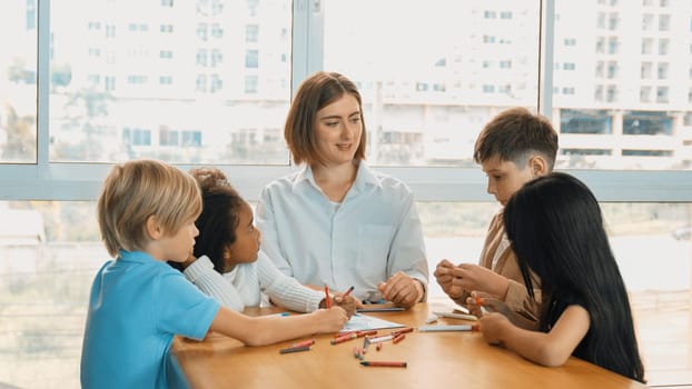 Professional caucasian teacher telling story to diverse student while sitting at table with storybook and colored book. Smart learner listening story while colored picture from instructor. Erudition.