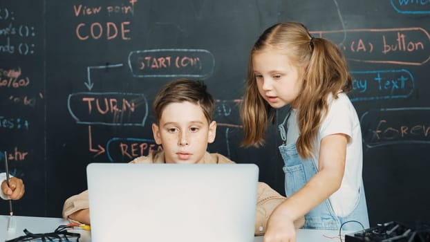Closeup of boy using laptop programing engineering code and writing program while group of diverse kid holding controller in STEM technology classroom at blackboard written with prompt. Erudition.