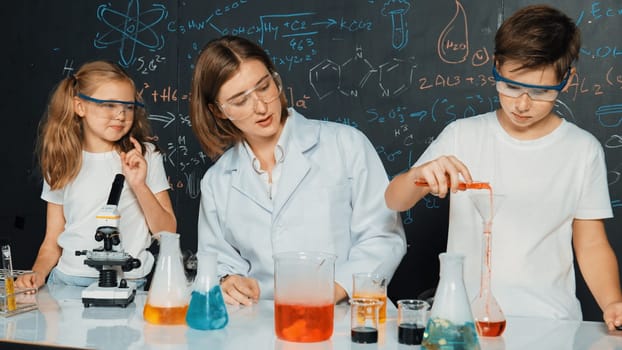 Caucasian boy mixing chemical liquid while teacher giving advice. Professional instructor wearing lab suit looking for diverse student at table with beaker filled with colored solution. Erudition.