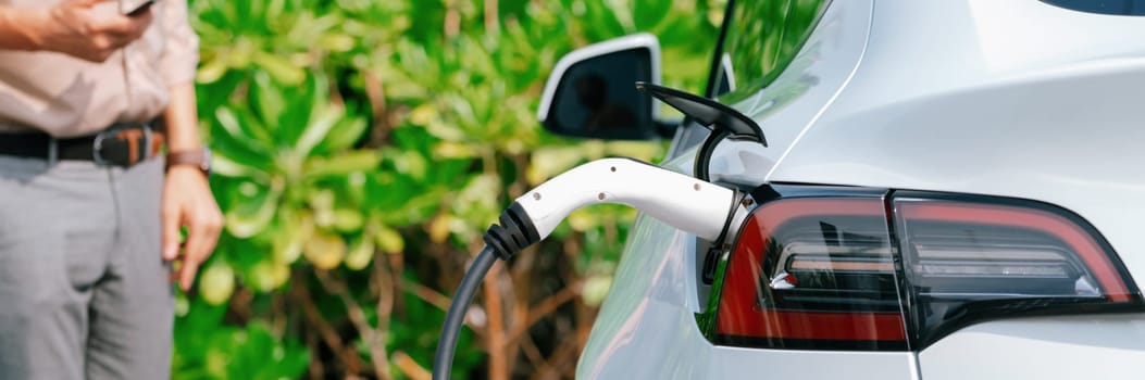 Young man use smartphone to pay for electricity at public EV car charging station green city park. Modern environmental and sustainable urban lifestyle with EV vehicle. Panorama Expedient