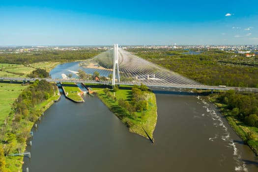 Cars drive on cable-stayed Redzinski Bridge over river flowing near scenic Wroclaw. Pylon bridge surrounded by lush green forests aerial motion along bridge