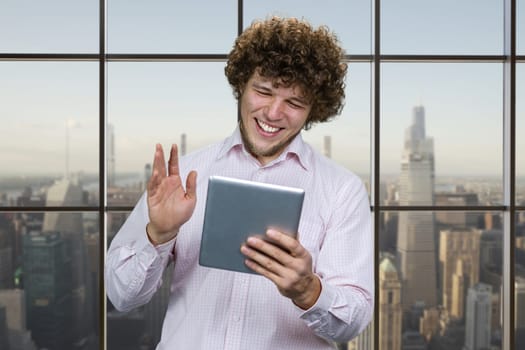 Portrait of a young happy caucasian man holding the tablet device and waving his hand. Checkered window with cityscape in the background.