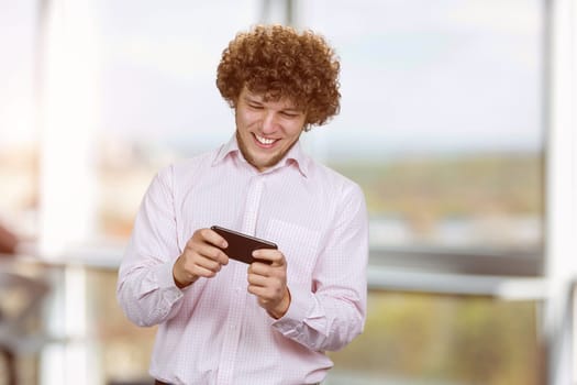 Excited happy young caucasian male gamer playing using smartphone. Guy playing mobile videogame app or watching digital video stream holding cell phone indoors.