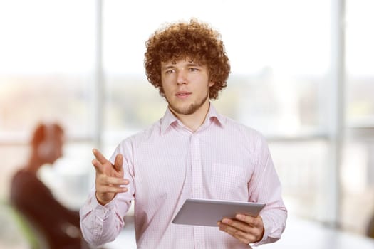 Portrait of a young businessman with curly hair standing with tablet pc device pointing at something. Office environment in the background.