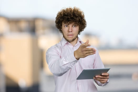 Portrait of a young caucasian worker with curly hair standing with tablet pc device pointing at something. Blurred urban scape in the background.