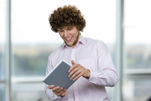 Portrait of a young happy caucasian man with curly hair playing a video game on the tablet pc device. Indoor window in the background.