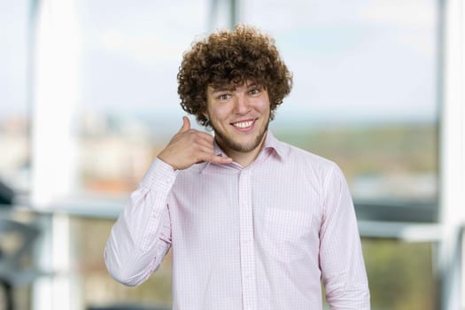 Handsome young man with curly hair shows call me gesture. Indoor window in the background.