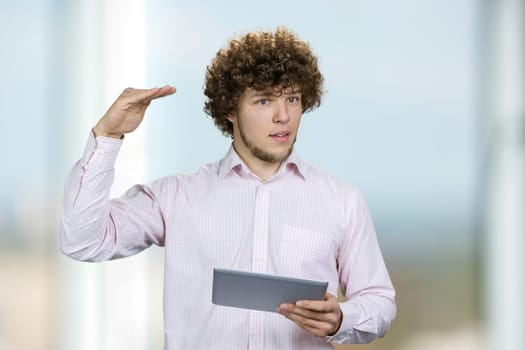 Portrait of a young man in white shirt holding a tablet pc showing height of something with hand. Male entrepreneur with curly hair giving a speech indoors.