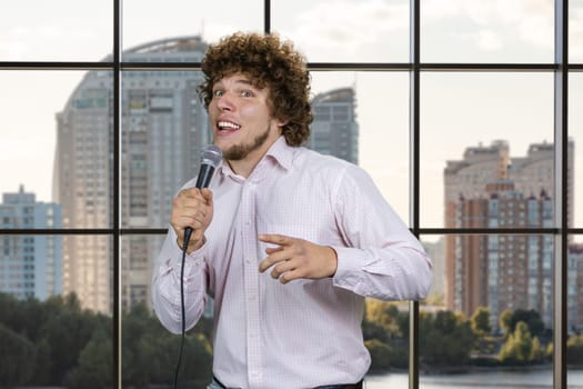 Happy excited young man with microphone standing indoors. Checkered window with residential area in the background.