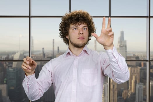 Portrait of a young man with curly hair and white shirt using invisible virtual screen for work. Indoor window background with cityscape view.