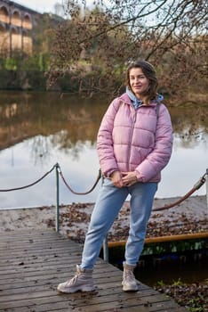 A young woman standing at the shore looking at the river in autumn sunny day. Street view, copy space for text, travel photo. Happy tourist woman on the bank of the river in autumn in warm clothes. Tourists enjoy their vacation, winter season. Romantic look and travel concept. A joyful mood in a Caucasian girl. Winter Wonderland: Enchanting Girl by the Riverside in Autumn.