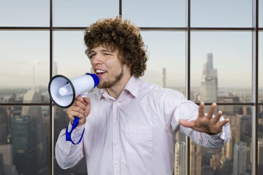 Portrait of a young excited man with curly hair in white shirt screaming hot news shout in megaphone. Indoor window in the background.