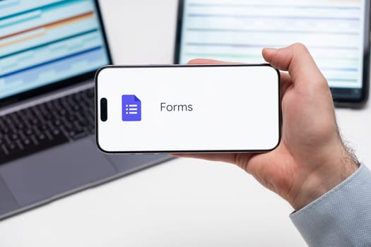 Forms application logo on the screen of smart phone in mans hand, laptop and tablet are on the table in the background, December 2023, Prague, Czech Republic.