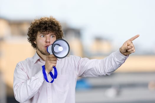 Young man with curly hair in white shirt speaks in megaphone pointing with index finger. Blurred urban scape in the background.