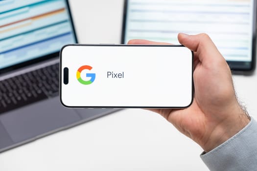 Google Pixel logo of app on the screen of mobile phone held by man in front of the laptop and tablet, December 2023, Prague, Czech Republic
