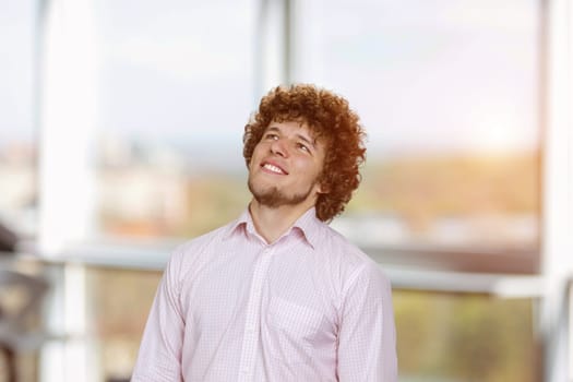Happy joyful smiling young man with curly hair looking up. Thinking of new good opportunities, dreaming. Indoor window in the background.