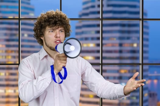 Handsome young man with curly hair in white shirt speaks in megaphone. Indoor window with night city view in the background.