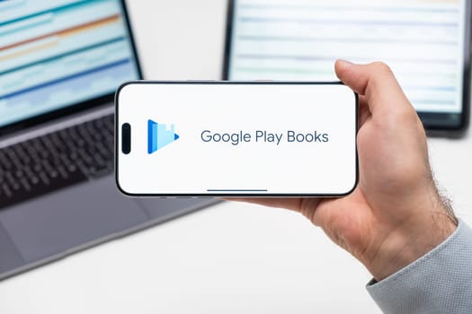 Google Play Books logo of app on the screen of mobile phone held by man in front of the laptop and tablet, December 2023, Prague, Czech Republic