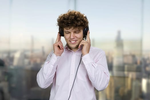 Happy young guy wearing headphones listening music. Blurred cityscape in the background.