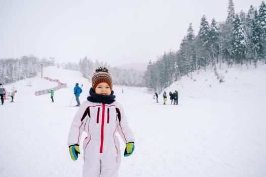 Little girl in a ski suit stands on a ski slope against the backdrop of skiers. High quality photo