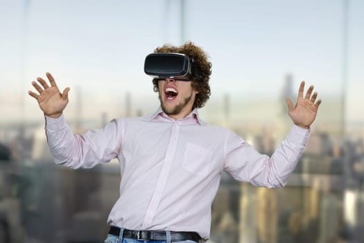 Portrait of young excited man with curly hair experiencing virtual reality. Blurred cityscape in the background.