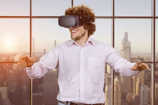 Portrait of happy young cheerful excited man with curly hair wearing vr headset. Indoor window with evening city view in the background.