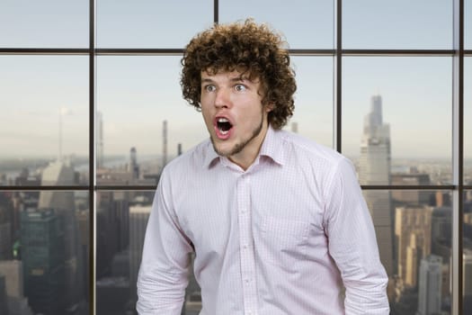 Young caucasian man with curly hair angrily screaming out loud. Checkered window with cityscape view in the background.