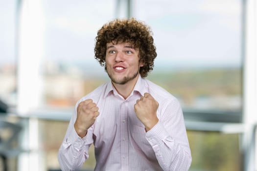 Portrait of happy excited young man with curly hair rejoicing success. Young football fan. Indoor window in the background.