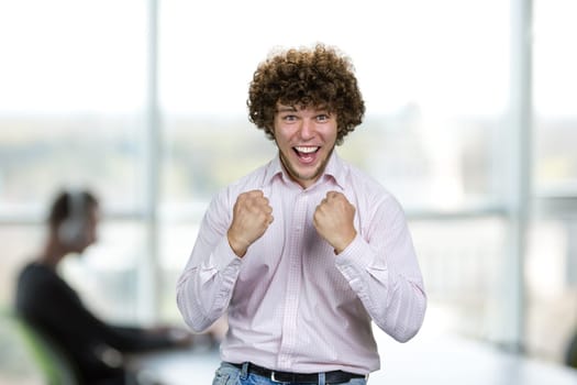 Portrait of happy excited young man with curly hair celebrates success. Indoor office interior in the background.