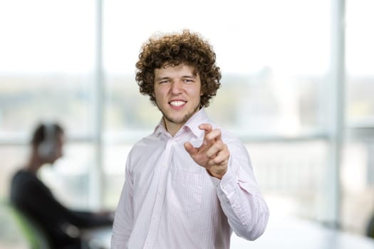 Portrait of a happy young cheerful man with curly hair shows his teeth and gesturing something. Blurred office interior in the background.