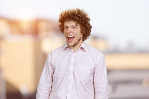 Portrait of a happy young man with curly hair with his mouth open. Blurred urban scape in the background.