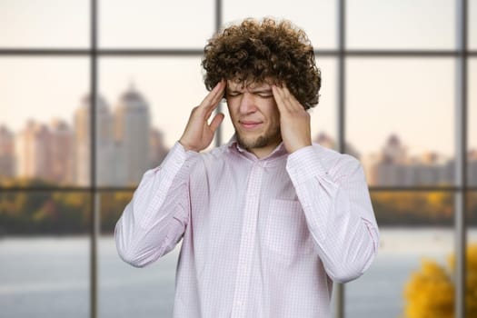 Young man in white shirt with curly hair having headache. Checkered window in the background.