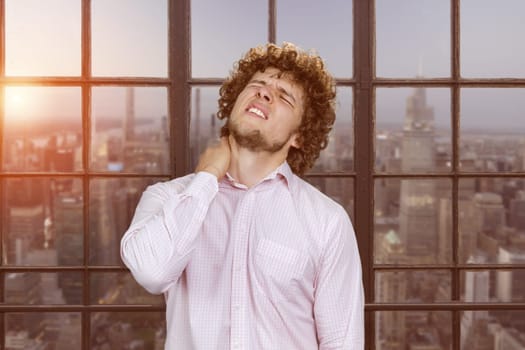 Portrait of a young caucasian man with curly hair having a neckache. Window with cityscape view in the background.