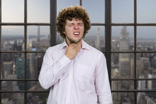 Young handsome man with curly hair suffers from neck pain. Indoor window with cityscape view in the background.